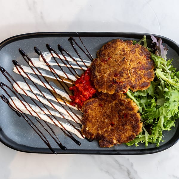 Two golden-brown crab cakes with red pepper relish, drizzled sauce, and fresh greens on a dark plate.