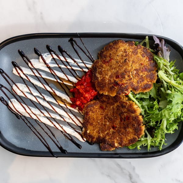 Two golden-brown crab cakes with red pepper relish, drizzled sauces, and a side of fresh greens.