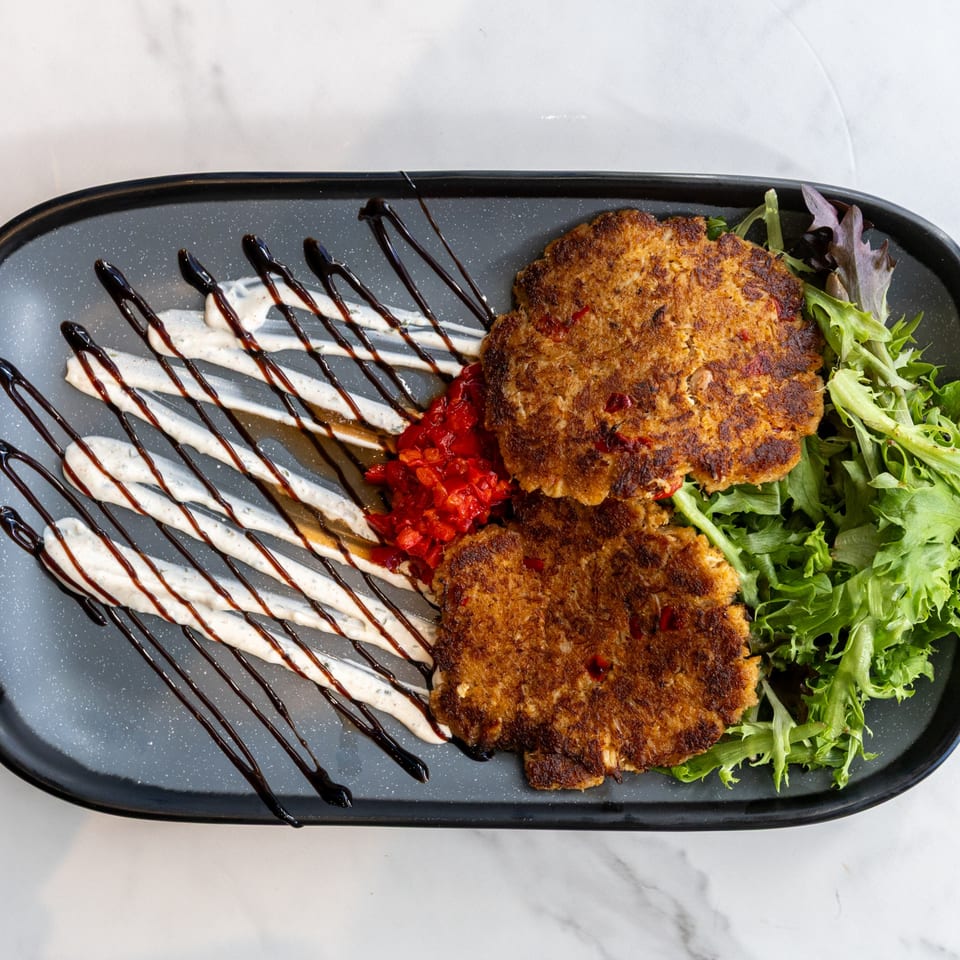 Two golden-brown crab cakes with red pepper relish, drizzled sauces, and a side of fresh greens.