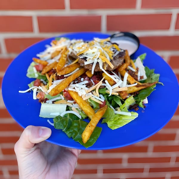 A hand holds a blue plate with a Dilworth Neighborhood Grille burger salad topped with fries and cheese.