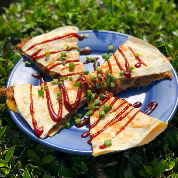 Four quesadilla wedges with barbecue sauce and green onions on a blue plate, surrounded by green foliage.