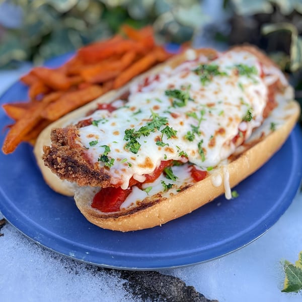 Chicken parmesan hero sandwich with melted cheese and parsley, served with sweet potato fries on a blue plate.