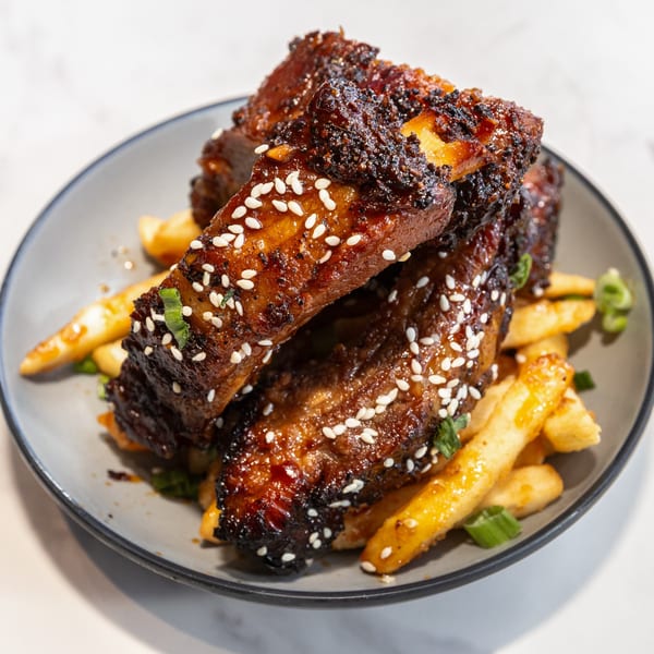 Glazed ribs with sesame seeds and green onions, served with golden french fries on a gray plate.