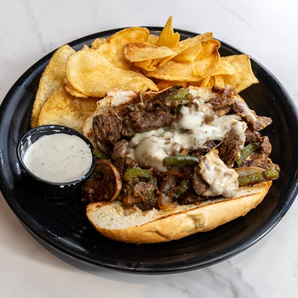 Cheesesteak sandwich with melted cheese, peppers, and onions, served with potato chips and ranch dip.