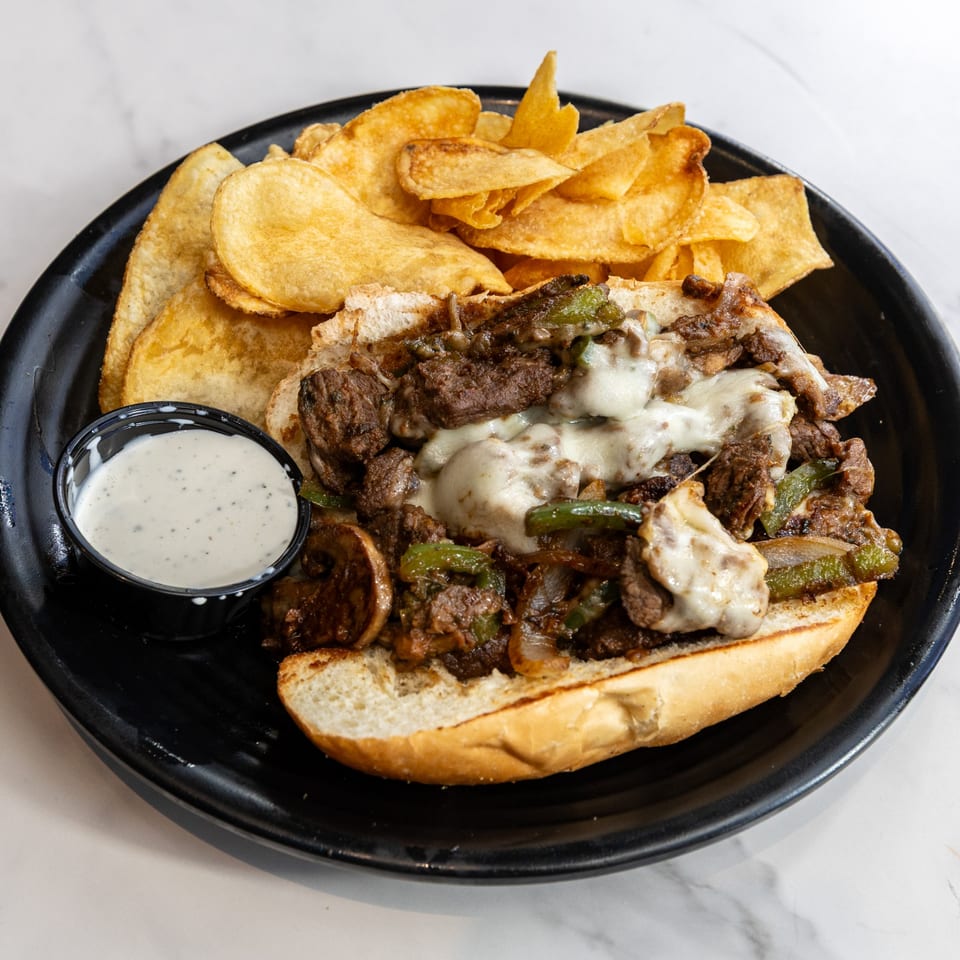 Cheesesteak sandwich with melted cheese, peppers, and onions, served with potato chips and ranch dip.