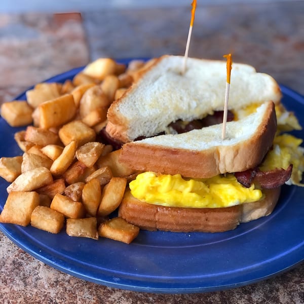Breakfast sandwich with scrambled eggs and bacon on toast, served with a side of home fries.